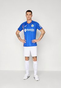 Modrý futbalový dres s oranžovými akcentmi a logami sponzorov, kombinovaný s bielymi nohavicami a ponožkami. Model stojí s rukami na bokoch.