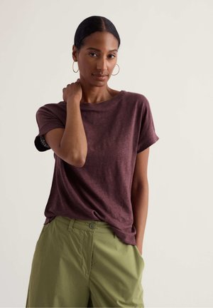 Femme aux cheveux foncés portant un t-shirt bordeaux à manches courtes et un pantalon vert clair, debout avec une main sur la nuque et l'autre dans la poche.