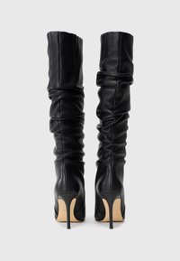 Schwarze, knielange High Heels mit schlaffem Lederdesign, von hinten auf einem schlichten, hellen Hintergrund betrachtet.