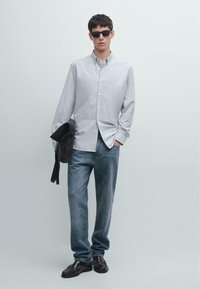 Koszula w paski z guzikami w jasnoszarym i białym kolorze, w połączeniu z niebieskimi dżinsami. Nosi czarne okulary przeciwsłoneczne i trzyma ciemną torbę. Czarne buty.