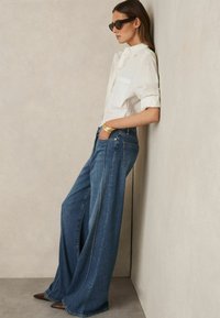 Jeans di denim a gamba larga con un lavaggio blu scuro, caratterizzati da cuciture bianche e un paio di mocassini. Abbinati a una camicia bianca e occhiali da sole.