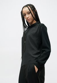 Zwarte capuchontrui met een losse pasvorm, lange mouwen en een getextureerde stof. Het ontwerp heeft een discreet zakje en contrasterende stiksels.