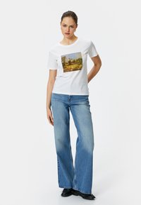 Camiseta de algodón blanca con un estampado artístico colorido en la parte frontal, combinada con unos pantalones de mezclilla de pierna ancha de color azul claro y zapatos negros.