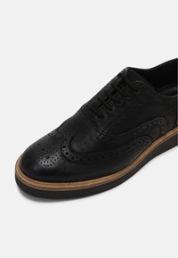 Chaussure brogues en cuir noir avec détails perforés, bout arrondi, fermeture à lacets et semelle en caoutchouc beige. Dispose d'une tige texturée et d'un design élégant.
