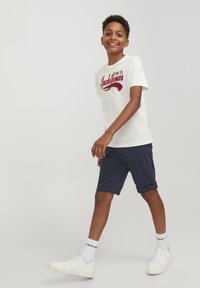 T-shirt bianca in cotone con stampa grafica rossa "Jack & Jones" sul davanti, abbinata a pantaloni blu scuro e sneakers bianche con calzini a costine.