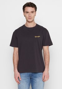 Camiseta de algodón negra con mangas cortas, escote redondo y logotipo de "Wrangler" bordado en amarillo en el pecho. Lleva puesto unos jeans de color azul claro.