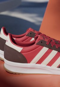 Paar rote Adidas-Sneaker mit weißen Streifen, brauner Ferse und weißer Cloudfoam-Sohle vor orange-blauem Hintergrund.