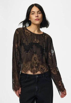 Femme aux cheveux noirs portant un haut en dentelle noire transparente à manches longues et un jean noir taille haute sur un fond blanc uni.
