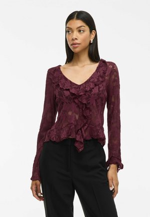 Blusa in pizzo borgogna con bordi arricciati, scollatura a V e maniche lunghe. Presenta un motivo testurizzato e un orlo inferiore svasato.