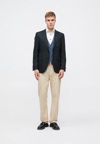 Sacou marin peste un veston albastru, cămașă albă și pantaloni bej. Material texturat, croială ajustată, cu pantofi negri slip-on.