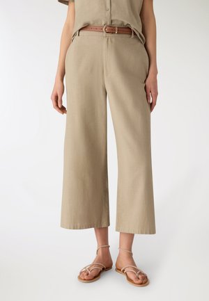Persona con pantaloni beige a gamba larga e taglio corto, cintura marrone e sandali piatti beige con cinturini, in piedi davanti a uno sfondo semplice.