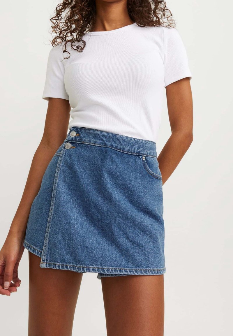 Femme portant un haut blanc à manches courtes avec une jupe asymétrique en jean bleu taille haute avec des détails de boutons.