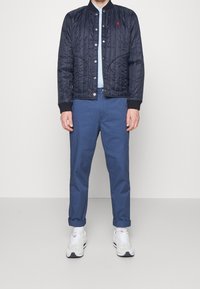 Marinblå vadderad bomberjacka med ribbad mudd, kombinerad med ljusblå skjorta och rullade marinblå chinos. Vita sneakers med grå detaljer.