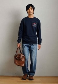 Sweat-shirt marine avec motif graphique, jean bleu clair, sac de sport en cuir marron et baskets marron. Tenue simple et décontractée avec une coupe ample.