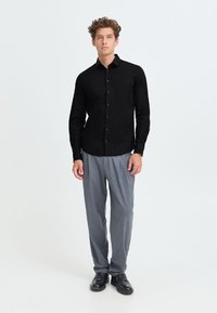 Giovane uomo in piedi con camicia nera abbottonata, pantaloni grigi e scarpe nere su sfondo bianco uniforme.