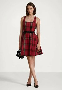 Γυναίκα με αμάνικο κόκκινο καρό φόρεμα και μαύρη ζώνη, κρατώντας μια μικρή μαύρη τσάντα και φορώντας μαύρες γόβες σε απλό φόντο.