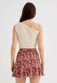 Top beige claro de un solo hombro con tejido texturizado, combinado con una falda roja acampanada que presenta un patrón de hojas blancas.