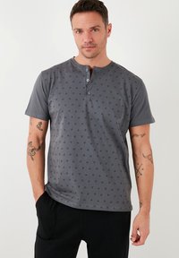 Camisa henley de manga corta gris con pequeños patrones de triángulos negros, que presenta dos botones en el cuello y una textura suave.
