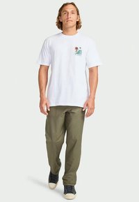 Man loopt naar voren en draagt een wit T-shirt met grafisch ontwerp, olijfgroene broek en zwarte sneakers tegen een effen lichte achtergrond.