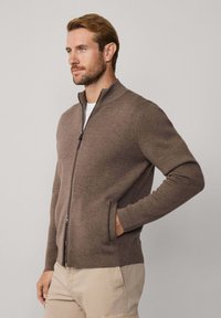 Hnedý kardigan na zips s vysokým golierom, vyrobený z textúrovanej látky. Má dlhé rukávy a bočné vrecká, nosený cez biele tričko.