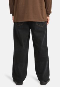 Pantaloni neri a gamba larga con tasche posteriori visibili, cuciture arancioni a contrasto e una texture liscia, abbinati a una maglietta marrone a maniche lunghe.