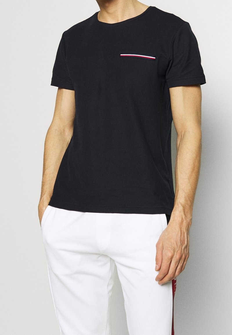 Schwarzes Baumwoll-T-Shirt mit kurzen Ärmeln, rundem Halsausschnitt und einer Brusttasche, die mit einem rot-weißen Streifenakzent verziert ist.
