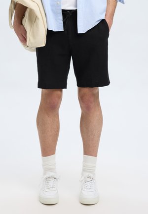 Bermudas negras hasta la rodilla con cintura ajustable mediante cordón. Combinadas con zapatillas blancas y calcetines cortos de color claro. El fondo es completamente blanco.