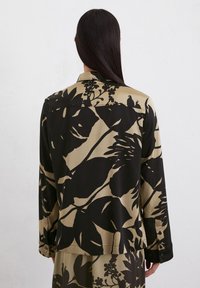 Chemise à manches longues avec un motif floral noir sur fond beige, fabriquée en tissu lisse, dotée d'un col classique et de poignets boutonnés.