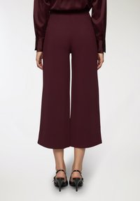 Pantaloni a gamba larga color bordeaux realizzati in tessuto morbido, caratterizzati da una vita alta e un fit rilassato, abbinati a tacchi neri con cinghie.