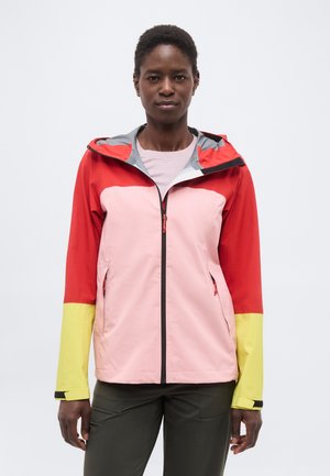 Γυναίκα που φορά ένα πολύχρωμο μπουφάν με κόκκινες, ροζ και κίτρινες περιοχές, ελαι πράσινο παντελόνι, στέκεται μπροστά σε ένα απλό λευκό φόντο.