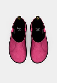Des chaussures roses à enfiler en matériau suédé doux, dotées de semelles en caoutchouc noires et de petites languettes en tissu au niveau du talon pour un enfilage facile.