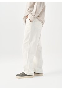 Personne debout, les mains dans les poches, portant un pantalon blanc ample, une chemise beige et des baskets grises avec des semelles blanches sur fond blanc.
