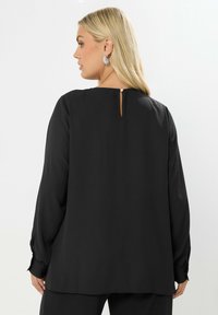 Blusa nera a maniche lunghe con dettaglio della schiena a fessura, texture liscia e polsini con bottoni. Vestibilità sagomata con silhouette rilassata.