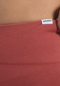 Ceinture de couleur rouille avec texture côtelée, au-dessus d'une zone de tissu lisse, avec une étiquette blanche affichant "SMILODOX" sur le côté.