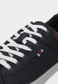 Κοντινό πλάνο μαύρου αθλητικού παπουτσιού με λευκά κορδόνια, ναυτικά λουριά και μικρή υφασμάτινη ετικέτα με ριγέ κόκκινο, λευκό και μπλε στο πλάι.