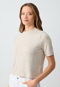 Top corto beige chiaro a maniche corte realizzato in un morbido tessuto testurizzato, con scollatura rotonda e vestibilità rilassata. Vista frontale con le mani sui fianchi.