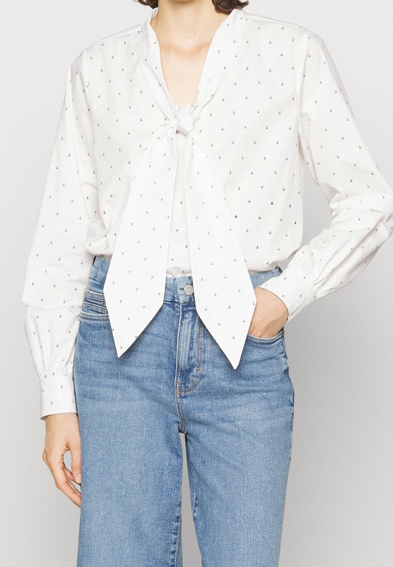 Blouse blanche à manches longues avec petits motifs d'ancre et un nœud devant, associée à un jean bleu taille haute, une main dans la poche.