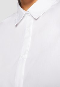 Gros plan d'une personne portant une chemise blanche impeccable à col pointu et trois boutons visibles fermés.