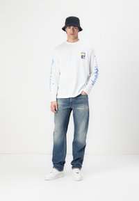 Cămașă albă cu mâneci lungi, având un grafic albastru pe mâneci, combinată cu blugi albaștri deteriorați și adidași albi. Modelul poartă o pălărie neagră de tip bucket.