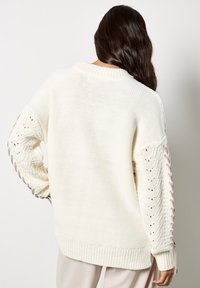 Kremowy sweter dzianinowy o luźnym kroju, z misternie zdobionymi rękawami w koronkowy wzór i ribbami na brzegach. Teksturowany materiał o miękkim wykończeniu.