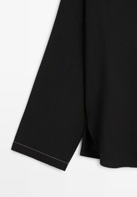 Chemise noire à manches longues avec un ourlet droit et une couture blanche visible près du poignet sur un fond blanc uni.