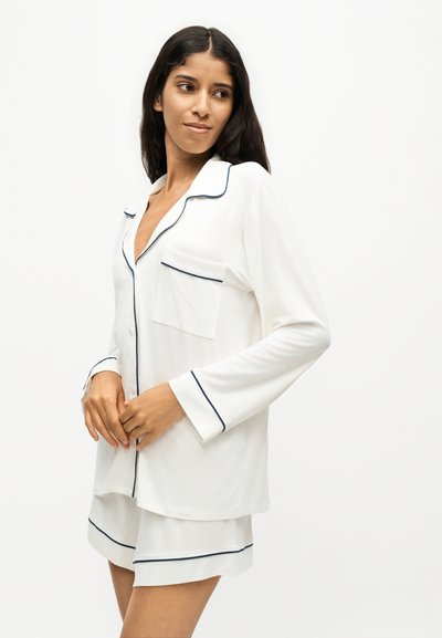 Femme portant un haut de pyjama blanc à manches longues et boutonné ainsi qu’un short assorti avec un liseré sombre, regardant sur le côté sur fond blanc.