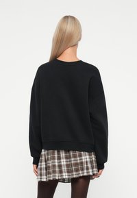 Mujer con pelo largo y rubio, vistiendo un sudadera negra de gran tamaño, falda de cuadros y pantys negros, de espaldas contra un fondo blanco.