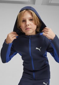 Sudadera con cremallera de color azul marino con detalles en azul, hecha de una tela suave. Cuenta con un bolsillo tipo canguro y un destacado logo blanco de Puma en el pecho.