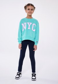 Felpa turchese con "NYC" in lettere rosa, abbinata a leggings blu scuro e sneakers bianche e nere. Tessuto morbido e vestibilità rilassata.