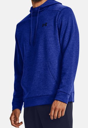 Man draagt een blauwe Under Armour hoodie met voorzak en zwarte broek tegen een effen achtergrond.
