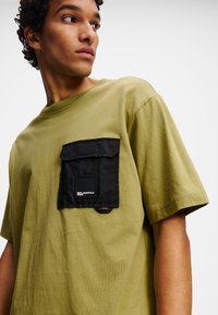 Alyvuogių žalios spalvos medvilninė marškinė su trumpomis rankovėmis ir juoda kišenė, kurioje yra logotipas. Lygus paviršius; laisvas siluetas.