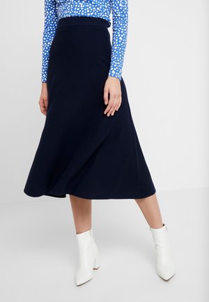 Femme portant une jupe midi marine, un haut bleu à manches longues à pois, et des bottines blanches, debout devant un fond uni.