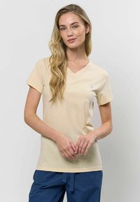 Világos bézs színű V nyakú póló, puha pamut anyagból, rövid ujjú, és egy diszkrét logó részlettel a bal mellen.