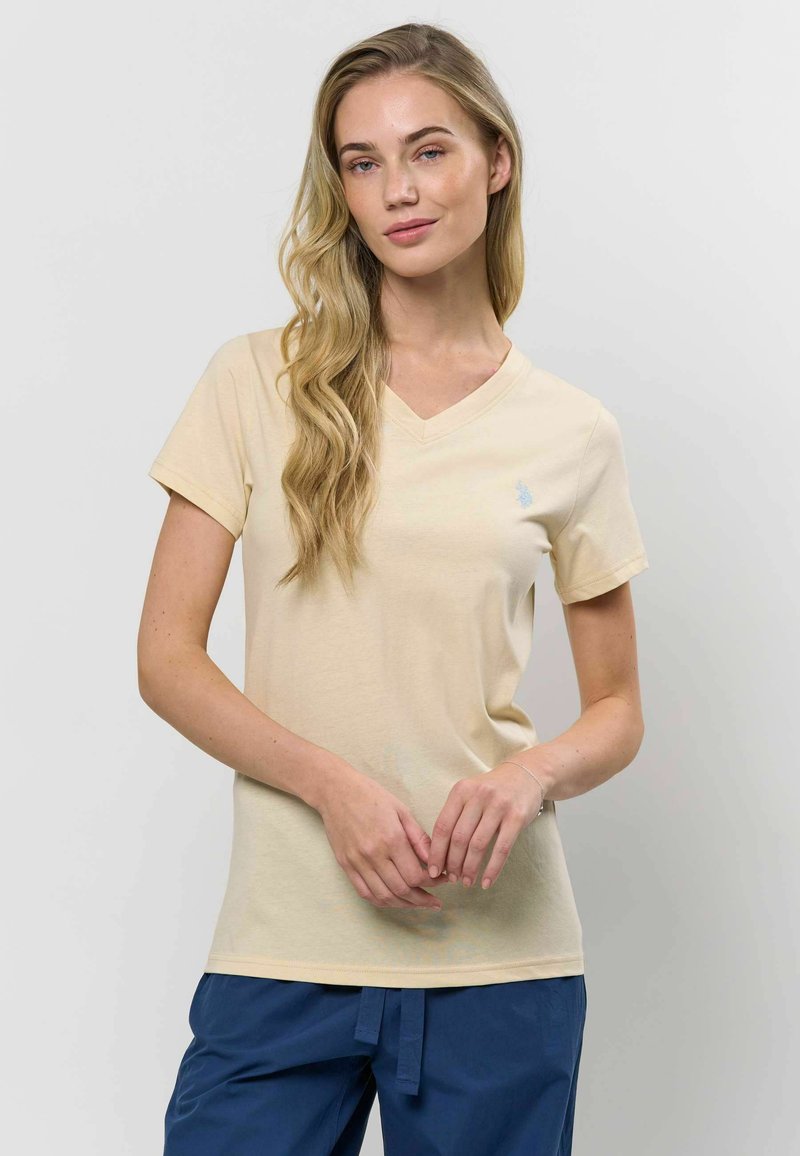 Šviesiai béžinės spalvos V formos marškinėliai, pagaminti iš minkštos medvilnės, su trumpomis rankovėmis ir nežymiu logotipo detaliu kairėje krūtinės pusėje.